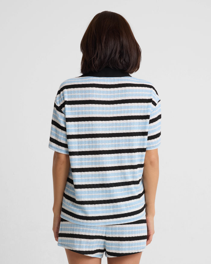 A person with shoulder-length dark hair stands with their back to the camera, wearing a relaxed fit Volcom Women's Coco Ho Rugby Top in light blue, black, and white stripes. The top features recycled polyester. Background is plain white.