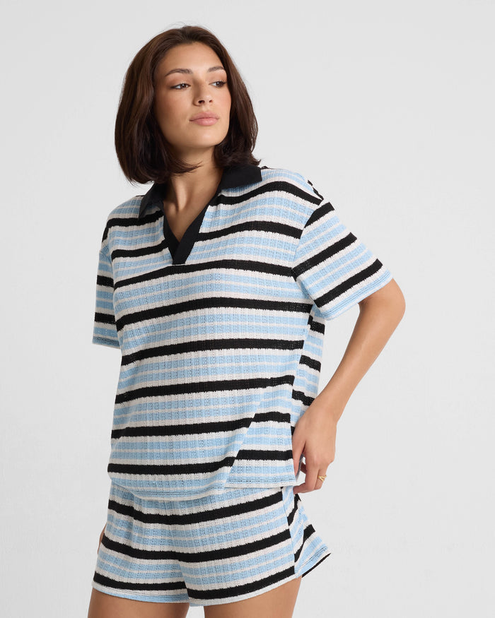 A woman with shoulder-length brown hair wears the Volcom Women's Coco Ho Rugby Top, a relaxed fit top with horizontal blue, black, and white stripes made from recycled polyester, paired with matching shorts, against a plain white background.