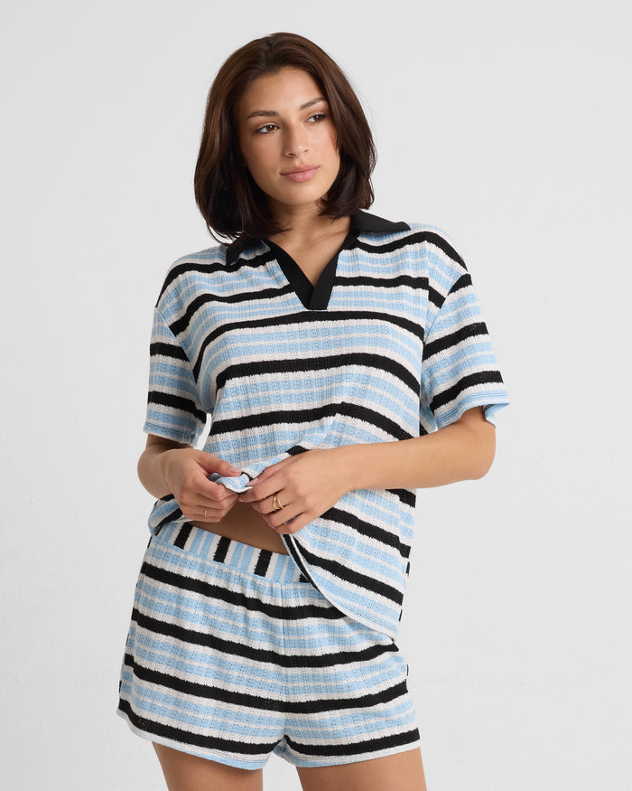 A woman with shoulder-length brown hair poses against a plain background, wearing a relaxed fit Volcom Women's Coco Ho Rugby Top in light blue, black, and white stripes. She pairs it with matching shorts and has a relaxed expression.