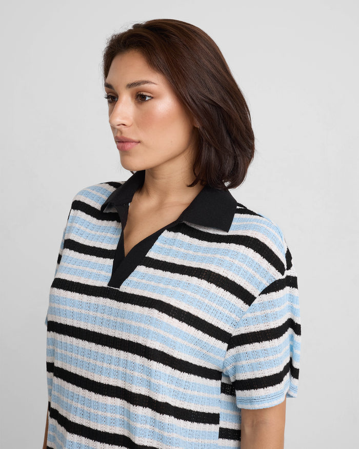 A woman with medium-length brown hair wears the Volcom Women's Coco Ho Rugby Top in blue, black, and white stripes, made from recycled polyester, standing against a plain light gray background.