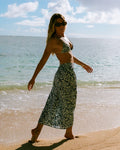 A woman in sunglasses, a bikini top, and the Volcom Women's Coco Ho Sandy Parts Skirt strolls barefoot on a sandy beach by the ocean under a partly cloudy sky.