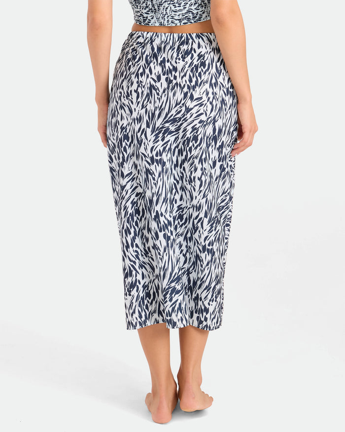 A person stands barefoot, facing away, wearing the Volcom Women's Coco Ho Sandy Parts Skirt—a high-waisted midi skirt with a black and white abstract pattern. The background is plain and light-colored.