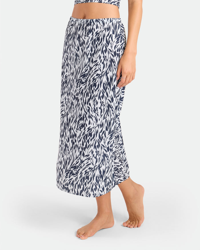A person stands barefoot in the Volcom Women's Coco Ho Sandy Parts Skirt, a high-waisted, ankle-length midi with a black and white abstract leaf print. The woven fabric drapes softly against a plain, light background.