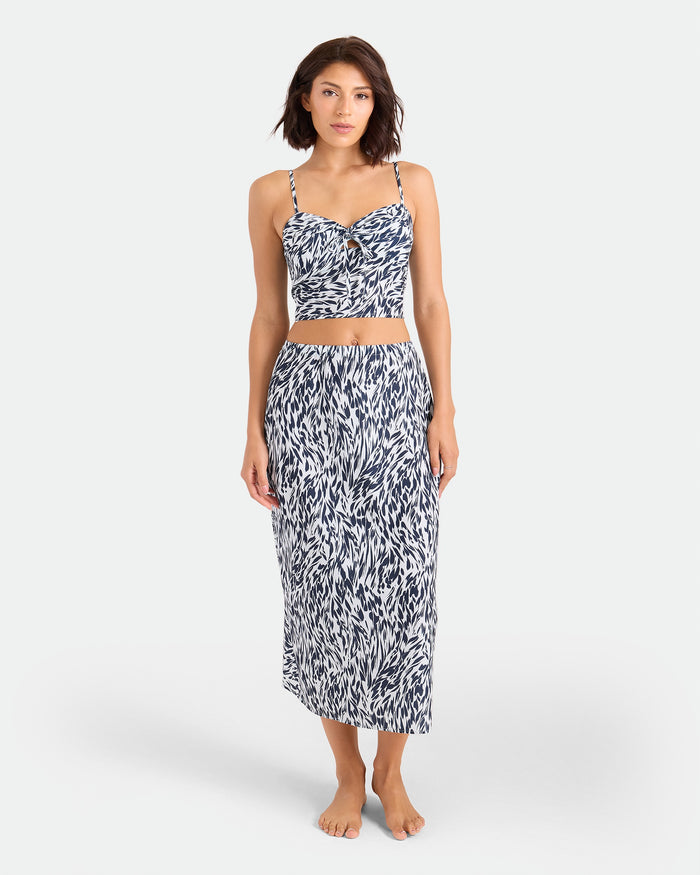 A woman stands barefoot in the Volcom Women's Coco Ho Sandy Parts Skirt set—featuring a fitted crop top and high-waisted, calf-length skirt with a blue and white abstract pattern—flowing elegantly against a plain white background.