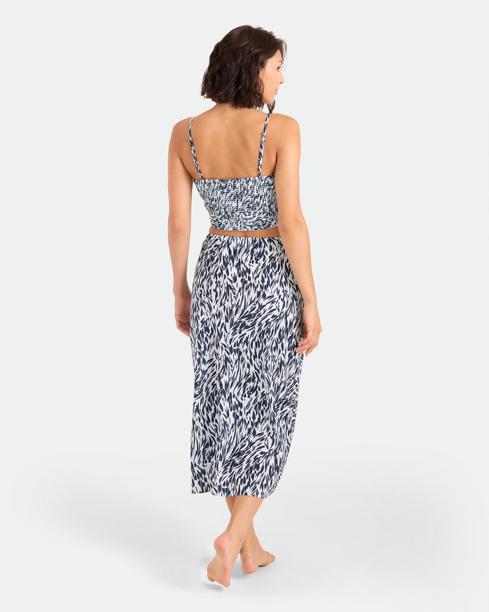 A woman with short brown hair stands barefoot, facing away. She wears the Volcom Women's Coco Ho Sandy Parts Skirt and a matching sleeveless crop top, both in blue with a soft woven drape, against a plain white background.