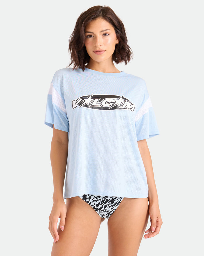 A woman with short brown hair wears a loose Volcom Women's Coco Ho Sports Jersey in the Summer Song colorway and black-and-white patterned bikini bottoms, standing against a plain light background.