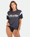 Coco Ho showcases the Women's Coco Ho Sports Jersey by Volcom—an oversized vintage black top with VOLCOM text—paired with black bikini bottoms, posing confidently against a plain light background.