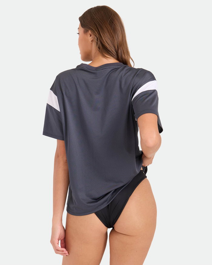 A woman with long brown hair faces away, wearing the Volcom Women's Coco Ho Sports Jersey in vintage black with white accents and black high-cut bottoms, set against a plain light background.