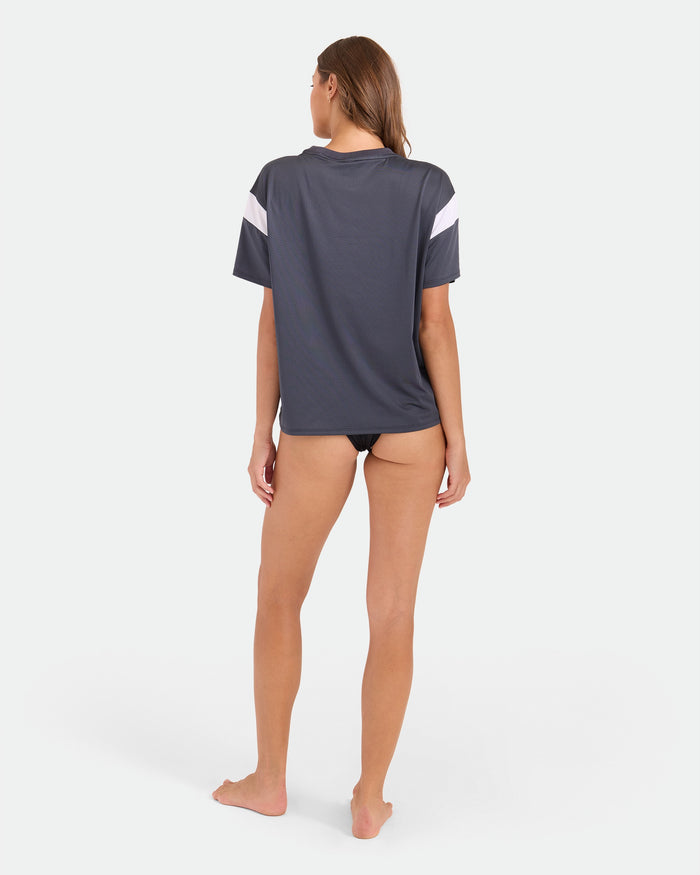 A woman stands barefoot on a white background, facing away from the camera. She wears the Volcom Women’s Coco Ho Sports Jersey in dark gray with white accents and vintage black bikini bottoms.