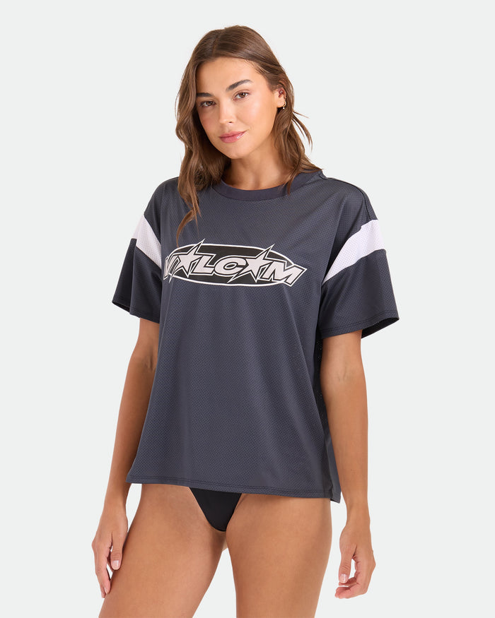 A woman wearing the Volcom Women's Coco Ho Sports Jersey, a vintage black oversized mesh T-shirt with white stripes and logo, paired with black bikini bottoms, poses against a plain light background.