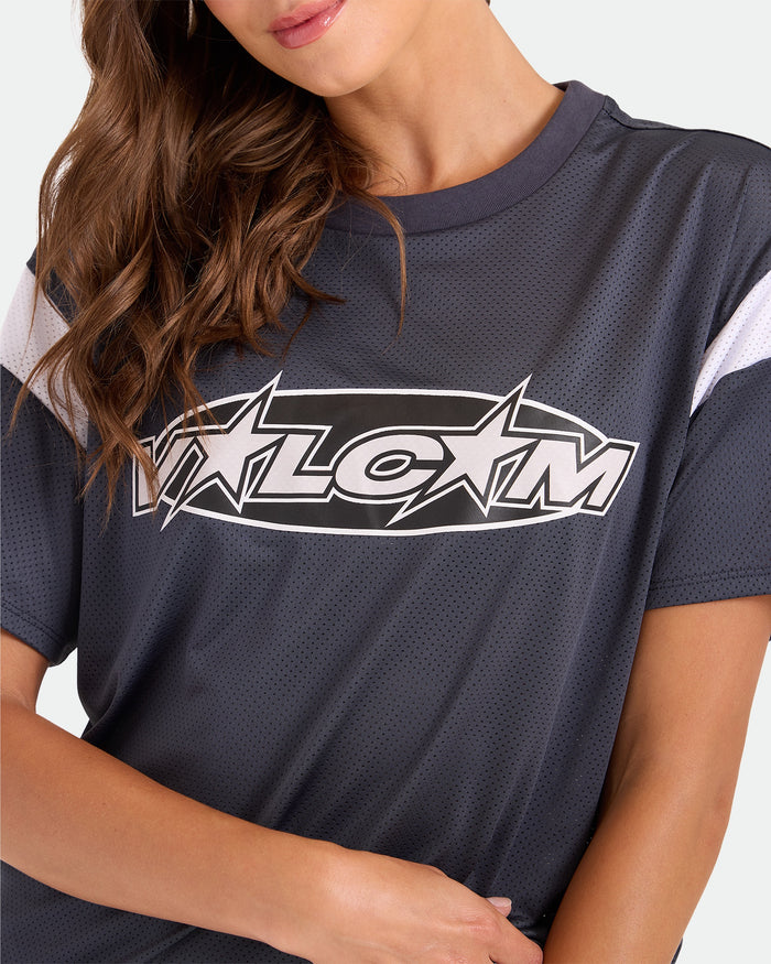 A woman with wavy brown hair wears the Volcom Women's Coco Ho Sports Jersey, a vintage black mesh shirt with a white VLCM logo and stars on the chest, posing with one arm across her body against a plain, light background.
