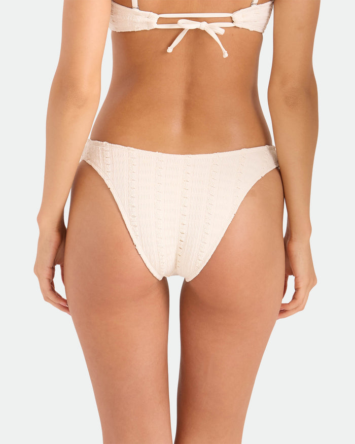 A woman is shown from the waist down wearing the Volcom Women's Coco Low Tide 90s V Bottom Skimpy bikini in cream, with textured fabric and minimal coverage, paired with a matching top, photographed from behind against a plain background.