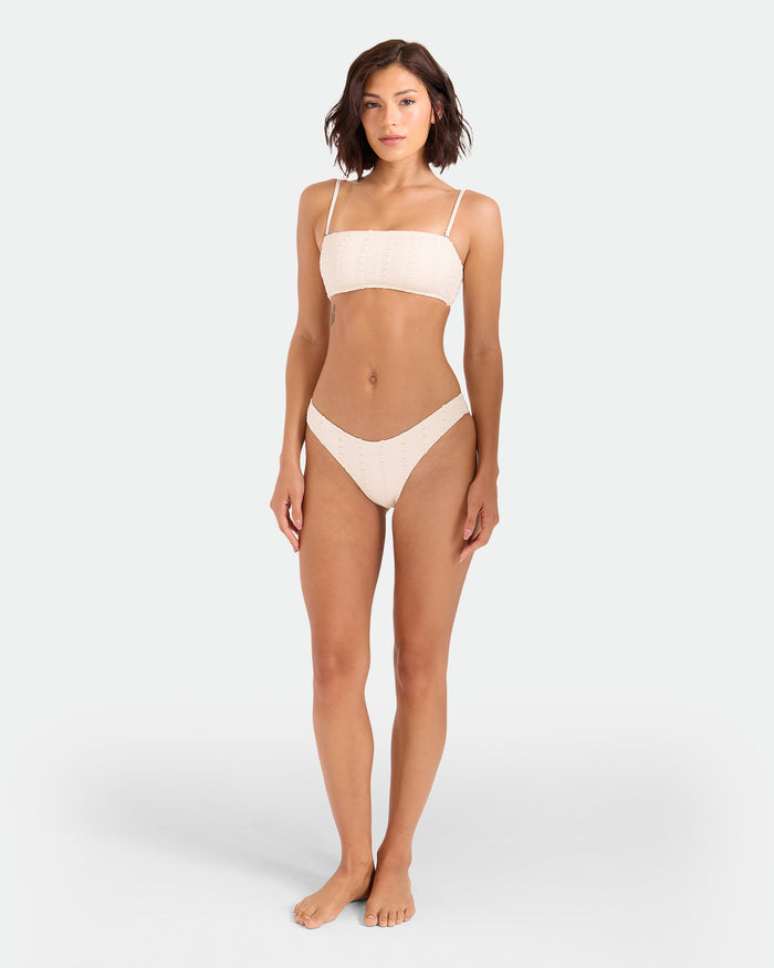 A woman with short brown hair stands barefoot against a plain light background, wearing the Volcom Women's Coco Low Tide 90s V Bottom Skimpy bikini, featuring a light-colored strapless top and matching high-leg cut bottoms.