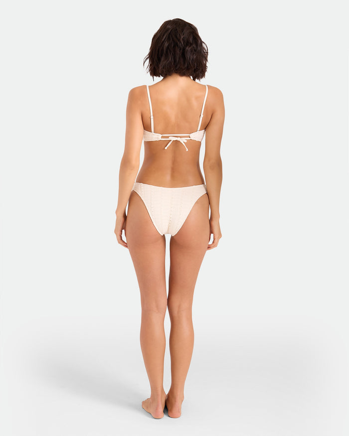 A woman stands barefoot facing away, wearing the Volcom Women's Coco Low Tide 90s V Bottom Skimpy bikini, featuring a tied back top and minimal coverage, set against a plain white background.
