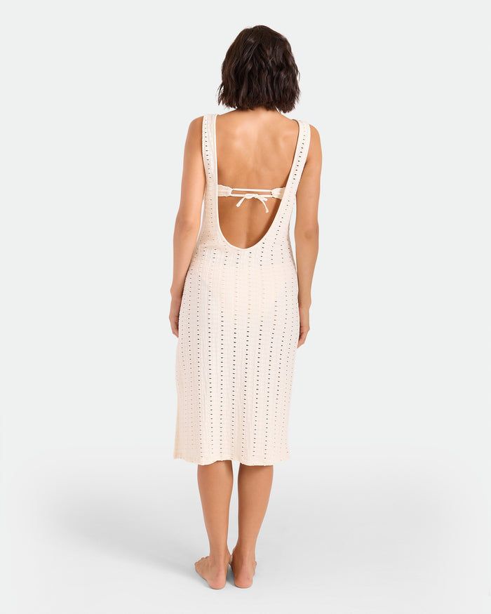 A woman stands barefoot facing away, wearing the Volcom Women's Coco Low Tide Midi Dress—a sleeveless, open-back knit body-con tank dress in light cream with a tie strap and below-the-knee length, set against a plain white background.