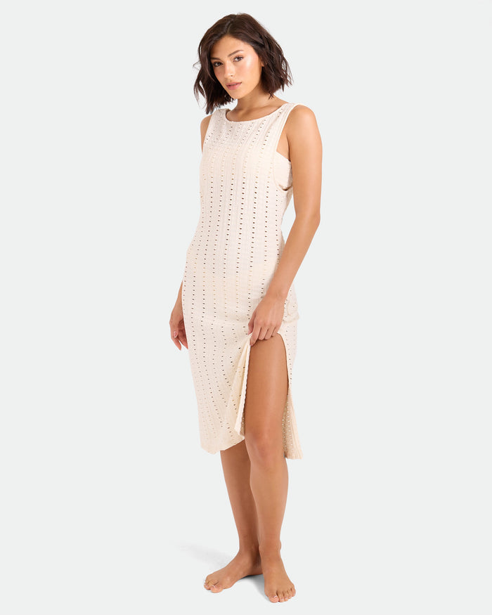 A woman stands barefoot in a sleeveless, light beige Volcom Women's Coco Low Tide Midi Dress with eyelet details and a high side slit, facing the camera with relaxed hands against a plain white background.