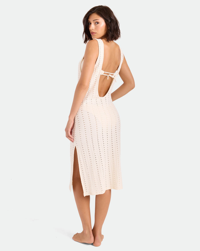 A woman with short dark hair models the Volcom Women's Coco Low Tide Midi Dress, a sleeveless, light-colored crochet tank dress with an open back and side slit, standing barefoot and glancing over her shoulder against a plain backdrop.