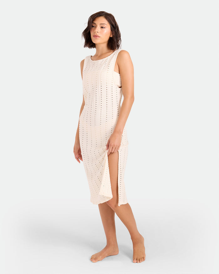 A woman with short brown hair wears the Volcom Women's Coco Low Tide Midi Dress—a sleeveless, light beige, perforated body-con tank dress with side slits—standing barefoot against a plain light background.