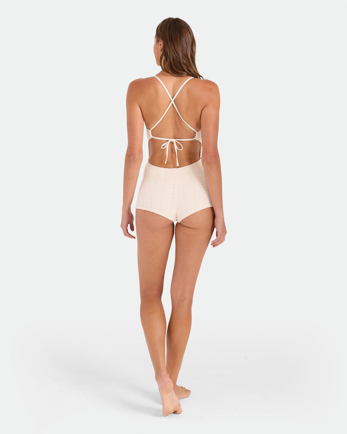 A woman with long brown hair stands barefoot, facing away, wearing the Volcom Women's Coco Low Tide Surf Suit One Piece in cream with a high waist and adjustable tie back straps, set against a plain white background.