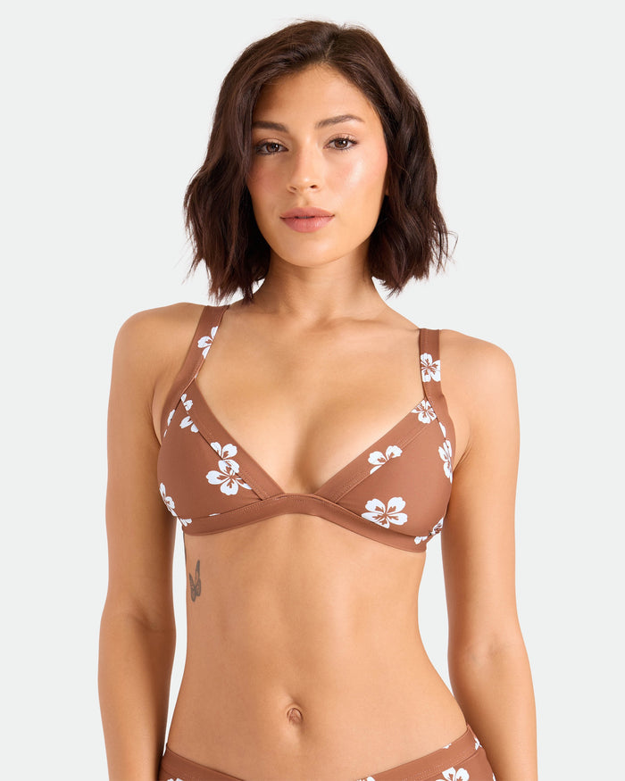 A woman with short brown hair wears the Volcom Women's Coco Retro Bloom Banded Triangle Top, a medium-support brown bikini with white floral patterns made from recycled nylon, standing against a plain light background.