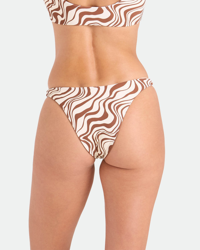 A person models the Volcom Women's Coco Retro Bloom Hipster Cheeky Reversible Bikini Bottom, featuring a brown and white wavy pattern, shown from the waist down with their back to the camera against a plain background.