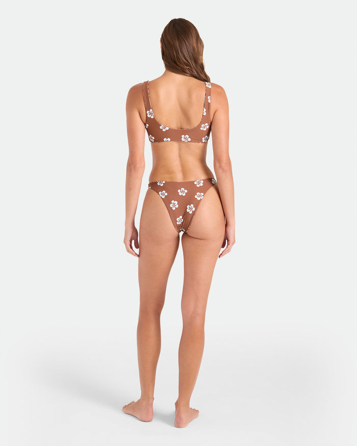 A woman stands barefoot, back to the camera, wearing the Volcom Women's Coco Retro Bloom Scoop Bralette Reversible with adjustable straps and white floral patterns, made from recycled nylon, set against a plain white background.
