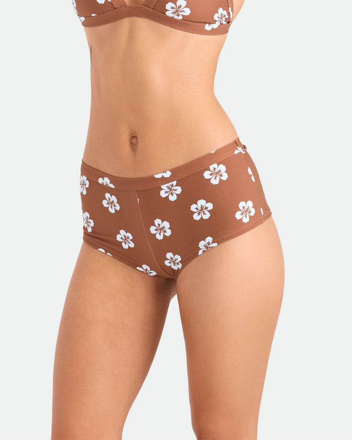 A model wears Volcom Women's Coco Retro Bloom Surf Shorts featuring a brown and white floral pattern for cheeky coverage, matching the bra, shown from chest to upper thighs against a plain light background.