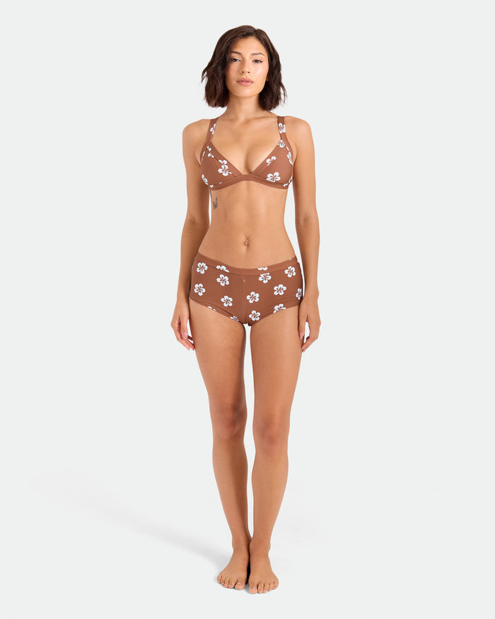 A woman stands barefoot against a white background, wearing Volcom's Women's Coco Retro Bloom Surf Shorts, a brown bikini bottom with white flower patterns made from recycled nylon, showcasing cheeky coverage.