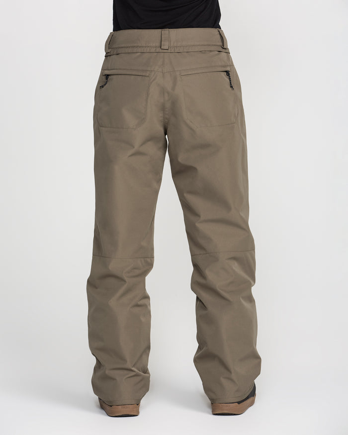 A person is shown from the back, wearing Volcom Women’s Coldfront Insulated Baggy Pants in a khaki color, featuring two zippered back pockets and belt loops, paired with brown shoes, all set against a plain white background.