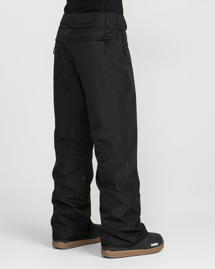 A person wearing Volcom Women’s Coldfront Insulated Baggy Pants in black, featuring V-Science 2-Layer construction, paired with black snow boots with brown soles, standing against a plain white background.