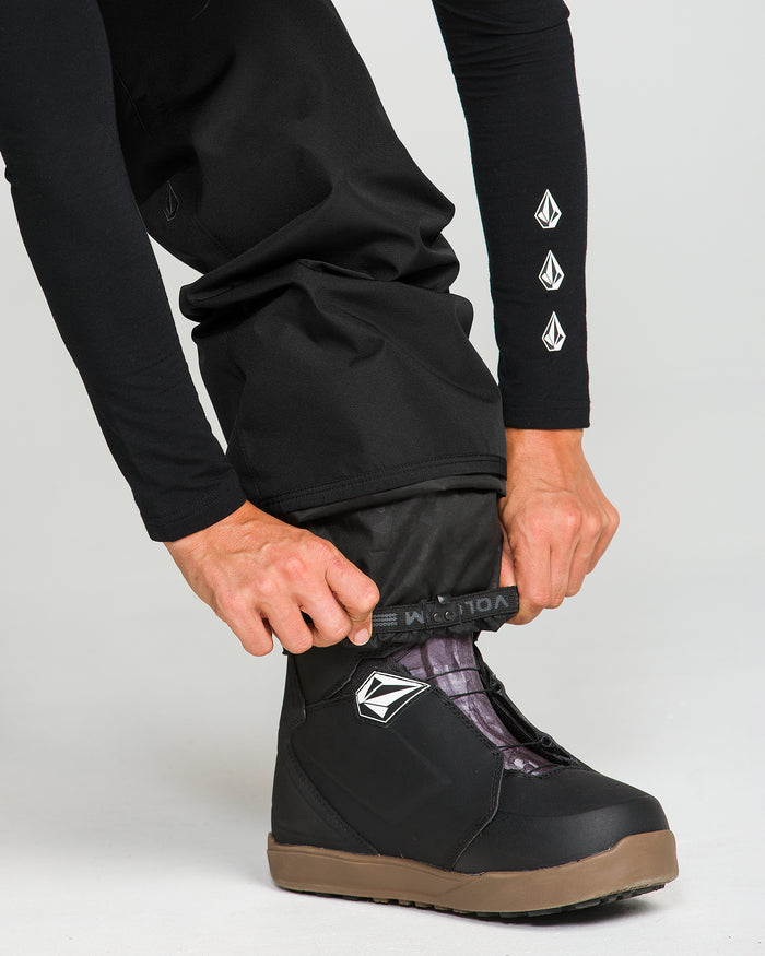 A person in black clothing adjusts a pair of Volcom Women’s Coldfront Insulated Baggy Pants—crafted with recycled insulation—over black and brown snow boots. The person’s shirt features three Volcom stone logos on the sleeve, matching the logo on both the pants and boots.