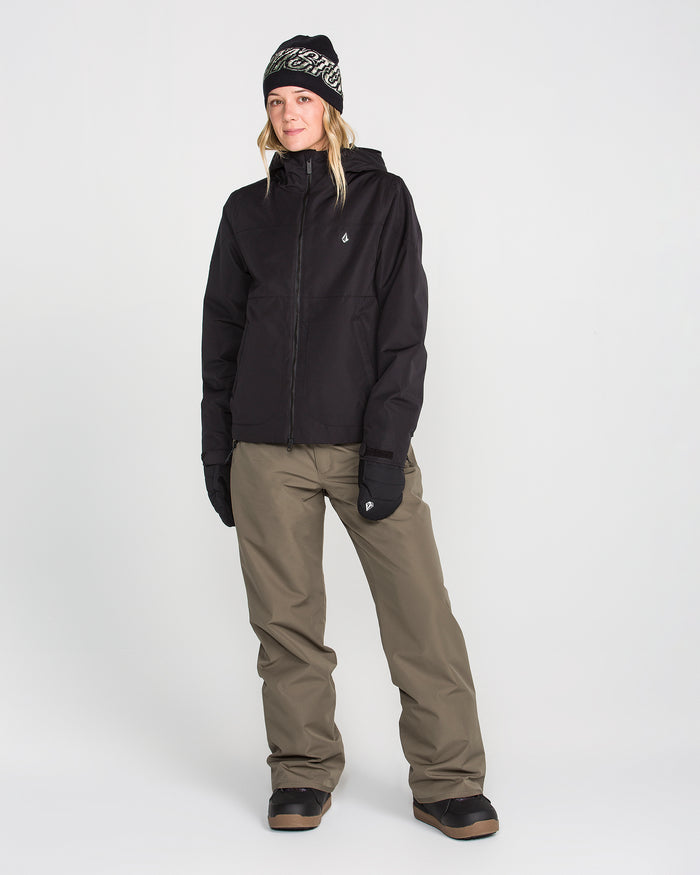 A person stands against a plain white background wearing black winter boots, black gloves, a black hooded jacket, a black beanie, and Volcom Women’s Coldfront Insulated Baggy Pants in olive green.