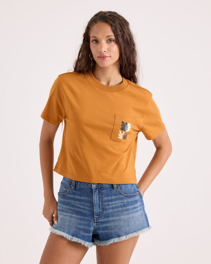 A woman models the Volcom Women's Core Memory Pocket Dial Tee—a cropped, mustard-hued top adorned with a subtle leaf design on the pocket—paired with frayed blue denim shorts. Made from 100% cotton, she stands against a simple, light backdrop.