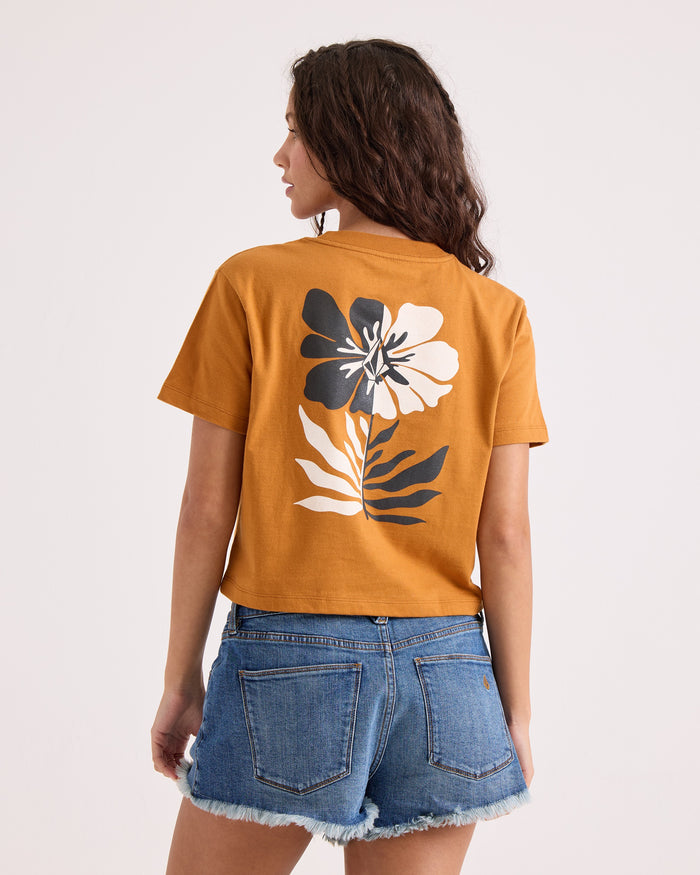 A woman with wavy brown hair wears the Volcom Women's Core Memory Pocket Dial Tee in mustard yellow—a 100% cotton, cropped silhouette with a large floral print on the back—styled with denim shorts and posed against a plain light background.