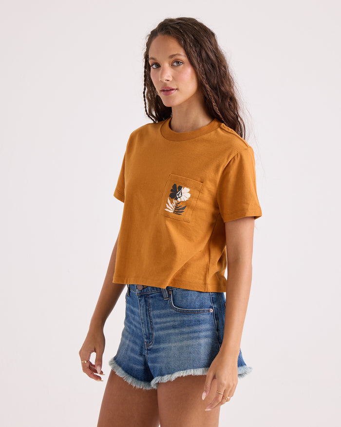 A woman wearing the Volcom Women's Core Memory Pocket Dial Tee—a 100% cotton mustard yellow t-shirt featuring a cropped silhouette and a floral pocket design—stands in denim shorts against a plain background, looking at the camera.