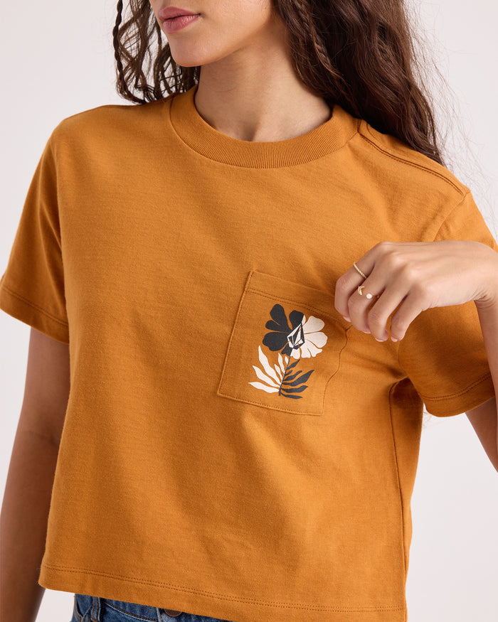 A woman wearing the Volcom Women's Core Memory Pocket Dial Tee in mustard yellow—a cropped, short-sleeve top made from 100% cotton with a floral-adorned front pocket—holds the pocket with her right hand.