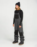 A person wearing a black beanie, a black long-sleeve shirt, and Volcom Women’s Creston 3D Stretch Bib overalls in gray with fully taped seams and black patches stands facing left against a plain white background.