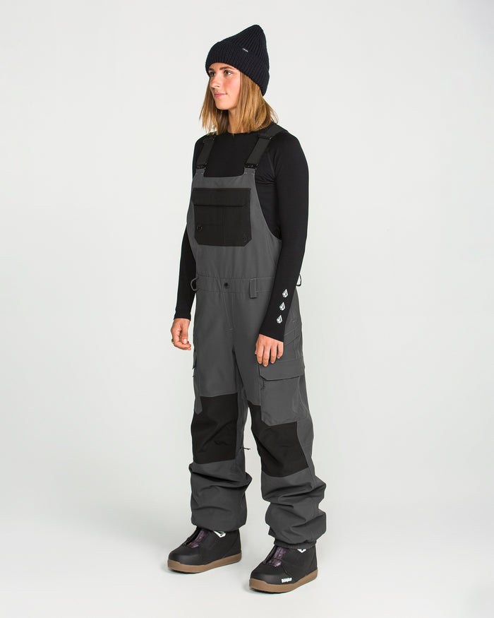 A person wearing a black beanie, a black long-sleeve shirt, and Volcom Women’s Creston 3D Stretch Bib overalls in gray with fully taped seams and black patches stands facing left against a plain white background.