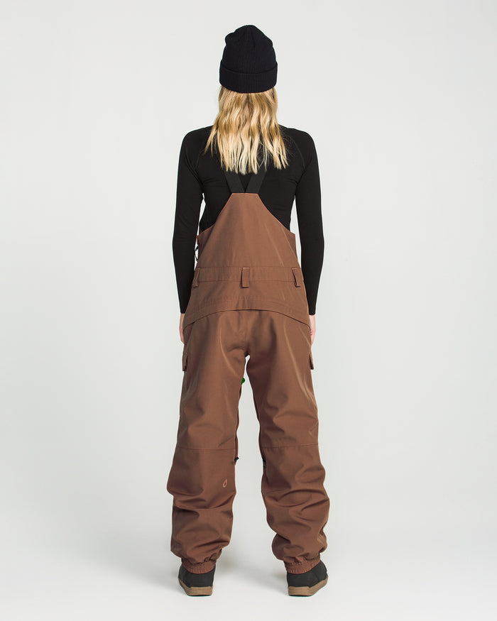 A person stands facing away against a plain white background, wearing a black beanie, a black long-sleeve top, and the Volcom Women’s Creston 3D Stretch Bib—brown wide-leg overalls crafted from recycled stretch oxford fabric.