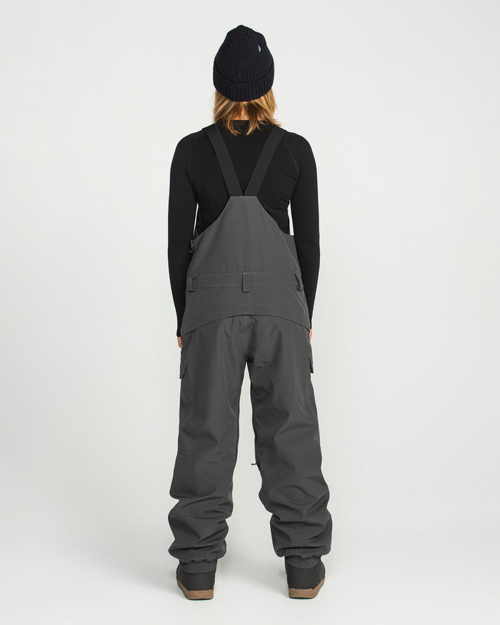 A woman stands facing away, dressed in a black beanie, black long-sleeve shirt, and Volcom Women’s Creston 3D Stretch Bib overalls in gray with wide legs and suspenders. The bib is crafted from recycled stretch oxford with fully taped seams, set against a plain white background.