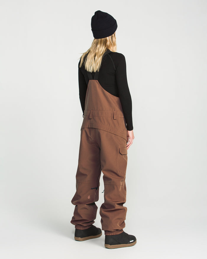 A person with long blonde hair wears the Volcom Women’s Creston 3D Stretch Bib in brown recycled stretch oxford fabric, a black beanie, and a black long-sleeve shirt. They are standing facing away from the camera against a plain white background.
