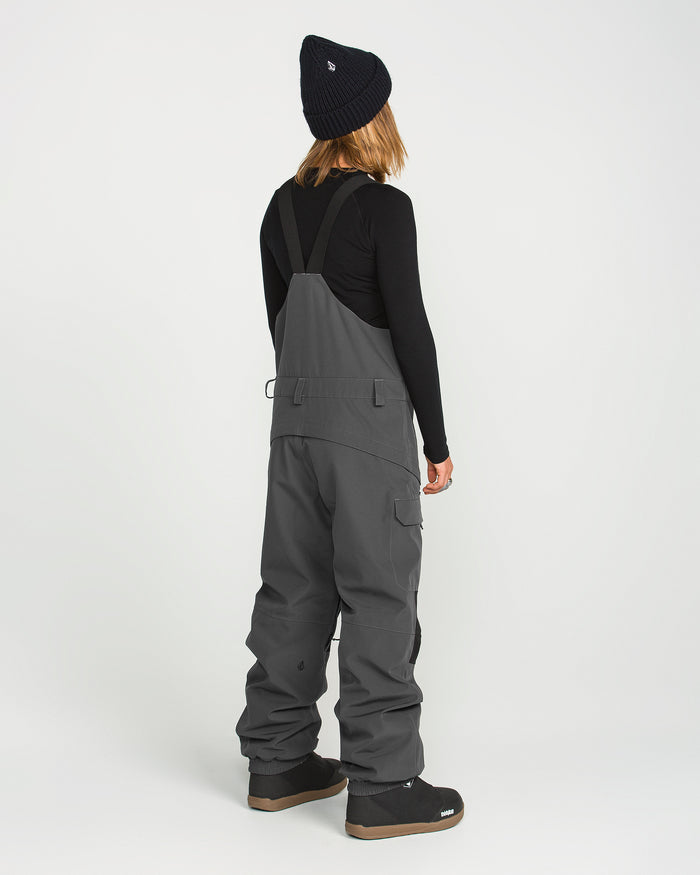 A person stands facing away, dressed in a black beanie, long-sleeve black shirt, and the Volcom Women’s Creston 3D Stretch Bib—crafted from recycled stretch oxford fabric with fully taped seams—in gray, paired with black boots against a plain white background.