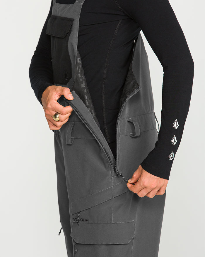 A person wearing a black long-sleeve shirt with white logos on the sleeve zips up the side of Volcom’s Women’s Creston 3D Stretch Bib in gray, showcasing its multiple pockets. The image highlights the torso and hand, drawing attention to the bib’s recycled stretch oxford fabric and fully taped seams for enhanced durability.