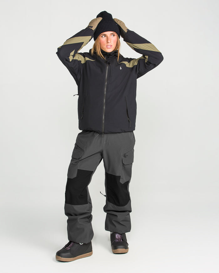 A person wearing the Volcom Women’s Creston 3D Stretch Bib—a black and beige jacket crafted from recycled stretch oxford—and a black beanie and gray cargo pants with fully taped seams stands with their hands on their head, looking to the side against a plain white background.
