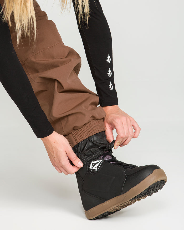 A person wearing Volcom’s Women’s Creston 3D Stretch Bib in brown, crafted from recycled stretch oxford fabric, and a black long-sleeve shirt adjusts the cuff of their bib pants over a black snow boot featuring a white geometric logo. The background is plain and light-colored.