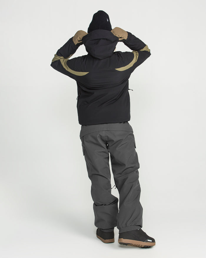 Wearing Volcom’s Women’s Creston 3D Stretch Bib, a person in winter clothing—including a black jacket with tan stripes made from recycled stretch oxford with fully taped seams, gray pants, brown gloves, black boots, and a black beanie—stands facing away and adjusts their hood.