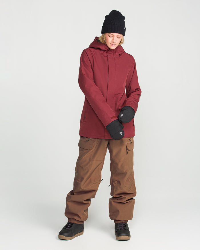 A person wearing a maroon jacket crafted from recycled stretch oxford, Volcom Women’s Creston 3D Stretch Bib snow pants in brown with fully taped seams, black hat, gloves, and boots stands against a plain white background, looking down with hands clasped together.