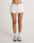 A person wears Volcom Women's Cruz Control vintage white high-rise denim shorts with a 5-pocket design, paired with white socks and sneakers, standing against a plain light background, legs shown from the waist down.