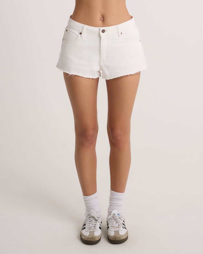 A person wears Volcom Women's Cruz Control vintage white high-rise denim shorts with a 5-pocket design, paired with white socks and sneakers, standing against a plain light background, legs shown from the waist down.