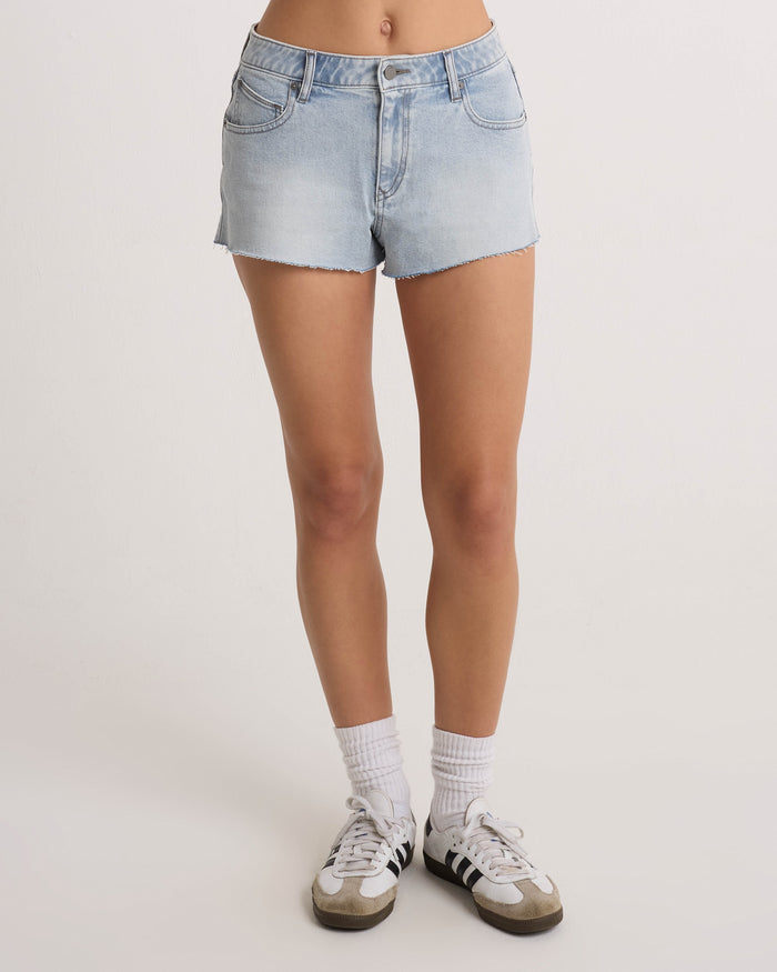 A person wears Volcom Women's Cruz Control high-rise denim shorts in chambray wash, paired with white socks and white sneakers with black stripes, standing against a plain light background. Only their legs and waist are visible.